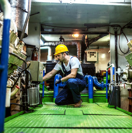 Mechanic in overalls and with helmet kneeling inside ship and repairing engine.